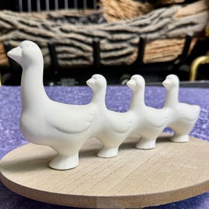 May include: A matte white ceramic figurine featuring a large adult goose followed by three smaller goslings in a single-file line. The sculpture is crafted from a smooth, unglazed material and is positioned on a circular, light-colored wooden base.