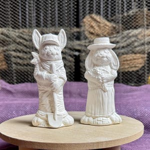 May include: Two white ceramic figurines of rabbits, one holding a shovel and the other holding a bouquet of flowers. The figurines are ready to be painted.