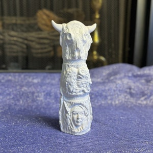 May include: A white, carved totem pole featuring a bison skull with horns, a bison, and a Native American face. The totem pole is standing upright on a purple surface.
