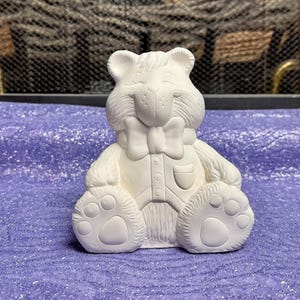 May include: A white ceramic bear figurine wearing a bow tie and a shirt with a pocket. The bear is sitting with its paws in front of it.