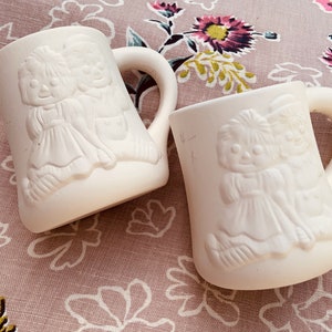 May include: Two white ceramic mugs with raised designs of two dolls. The dolls are wearing dresses and have long hair. The mugs have handles.