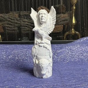 May include: A white ceramic sculpture depicting a Native American figure with an eagle and a buffalo. The sculpture features detailed carvings of feathers, a headdress, and the word "SEE". The statue is on a blue surface.
