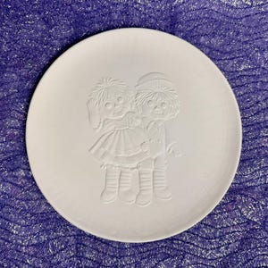 May include: White ceramic plate with a raised design of two dolls holding hands. The dolls are wearing simple dresses and hats.