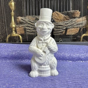 May include: A white ceramic bear figurine wearing a top hat, bow tie, and vest, holding a guitar. The bear stands on a barrel-shaped base. The figurine is set against a purple surface and a fireplace screen.