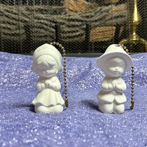May include: Two white ceramic figurines of children with their hands clasped in prayer. The figurines are attached to gold chains, which are likely used as light pull chains.