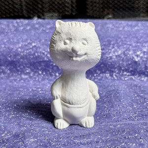 May include: A white ceramic figurine of a smiling cartoon beaver. The beaver has a textured fur appearance, small ears, and is standing upright. It is wearing a simple garment around its waist. The figurine is on a purple surface.