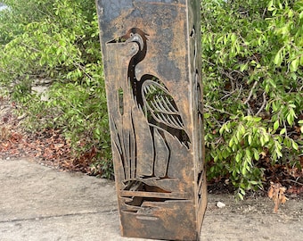 May include: A tall, rectangular metal garden sculpture with a weathered, rusted finish. The front panel features a laser-cut silhouette of a heron standing among cattails and water reeds, creating a detailed cutout design on the dark metal surface.