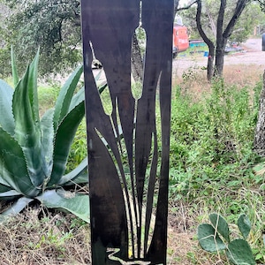 May include: A metal garden sculpture with a cut-out design of tall grass and reeds. The sculpture is painted a dark brown color and is standing in a grassy area.