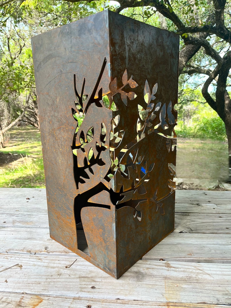 May include: A rusty metal planter with a cut-out design of a tree with leaves. The planter is square and has a rustic look.