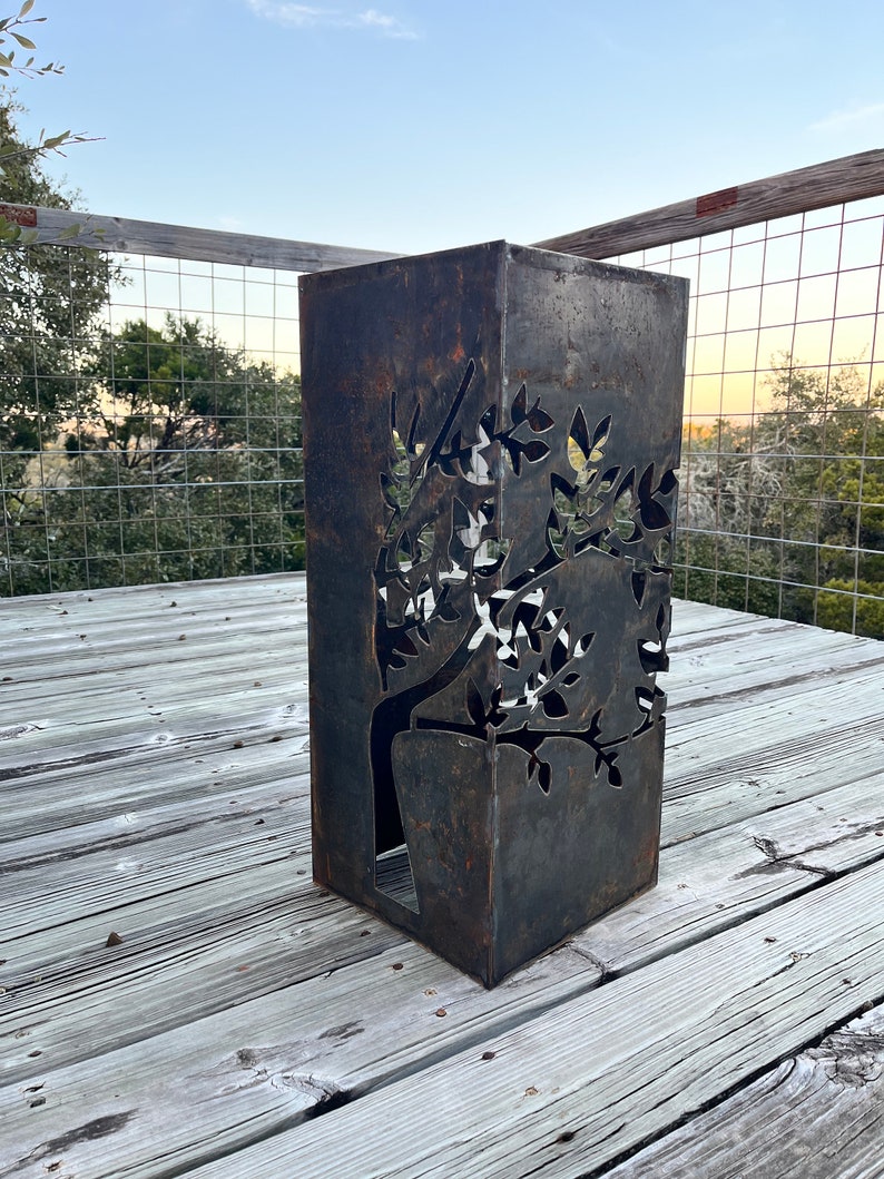May include: A rusty metal planter with a tree design cut out. The planter is on a wooden deck.