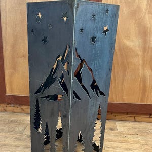 May include: A dark metal lantern with cut-out mountain, star, and tree designs. The lantern is rectangular and stands on a wooden floor. The cut-out designs allow light to shine through, creating a decorative effect.