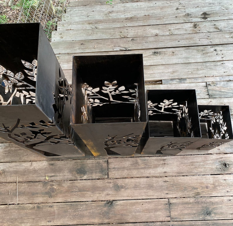 May include: Four black metal planters with a tree design cut out. The planters are stacked on top of each other, with the largest planter at the bottom and the smallest planter at the top.
