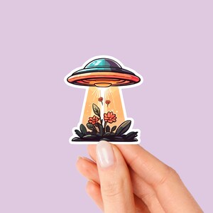 Könnte beinhalten: Eine Cartoon-Illustration eines fliegenden Untertassers, der über einem kleinen Stück grünem Gras mit roten Blumen schwebt. Die Untertasse strahlt einen Lichtstrahl aus.