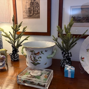 Könnte beinhalten: Eine dekorative Anordnung mit einem weißen Porzellan-Pflanzgefäß mit Vogel- und Schmetterlingsillustrationen, flankiert von kleinen immergrünen Bäumen, die in schwarz-weiß kariertem Stoff verpackt sind. Eine Glasbox, eine kleine blaue Geschenkbox und ein dekorativer Teller sind ebenfalls vorhanden.