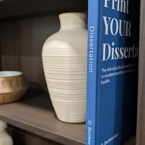 Puede incluir: Un libro azul titulado "Print YOUR Dissertation" con texto blanco, de pie sobre una estanter&iacute;a de madera. El lomo del libro dice "Dissertation" verticalmente. El libro trata sobre la inteligencia artificial en la atenci&oacute;n m&eacute;dica. Otros elementos decorativos son visibles.