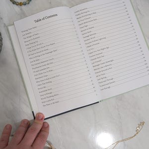 Pu&ograve; includere: Un libro aperto con un sommario, con titoli come "Wishing Time Away" e "Just Breathe". Sono visibili anche un braccialetto di perline e una collana d'oro. Il libro &egrave; su una superficie chiara.