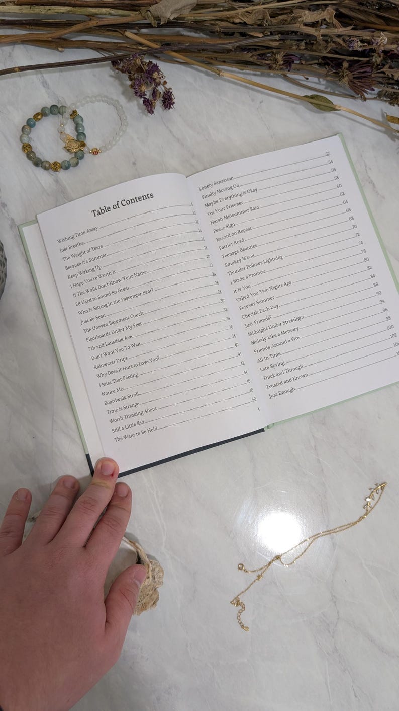 Pu&ograve; includere: Un libro aperto con un sommario, con titoli come "Wishing Time Away" e "Just Breathe". Sono visibili anche un braccialetto di perline e una collana d'oro. Il libro &egrave; su una superficie chiara.