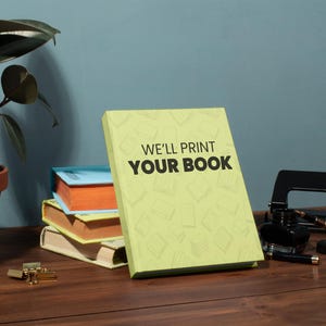 May include: A stack of books with a light green book in the front that says "WE'LL PRINT YOUR BOOK". The books are on a wooden surface with a potted plant, inkwell, and hole puncher.