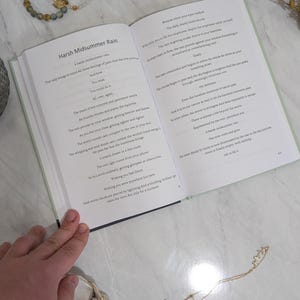 Pu&ograve; includere: Un libro aperto con il titolo "Harsh Midsummer Rain" e testo sulle pagine. Due braccialetti di perline e una collana d'oro sono accanto al libro. Fiori e steli secchi sono sparsi intorno al libro.