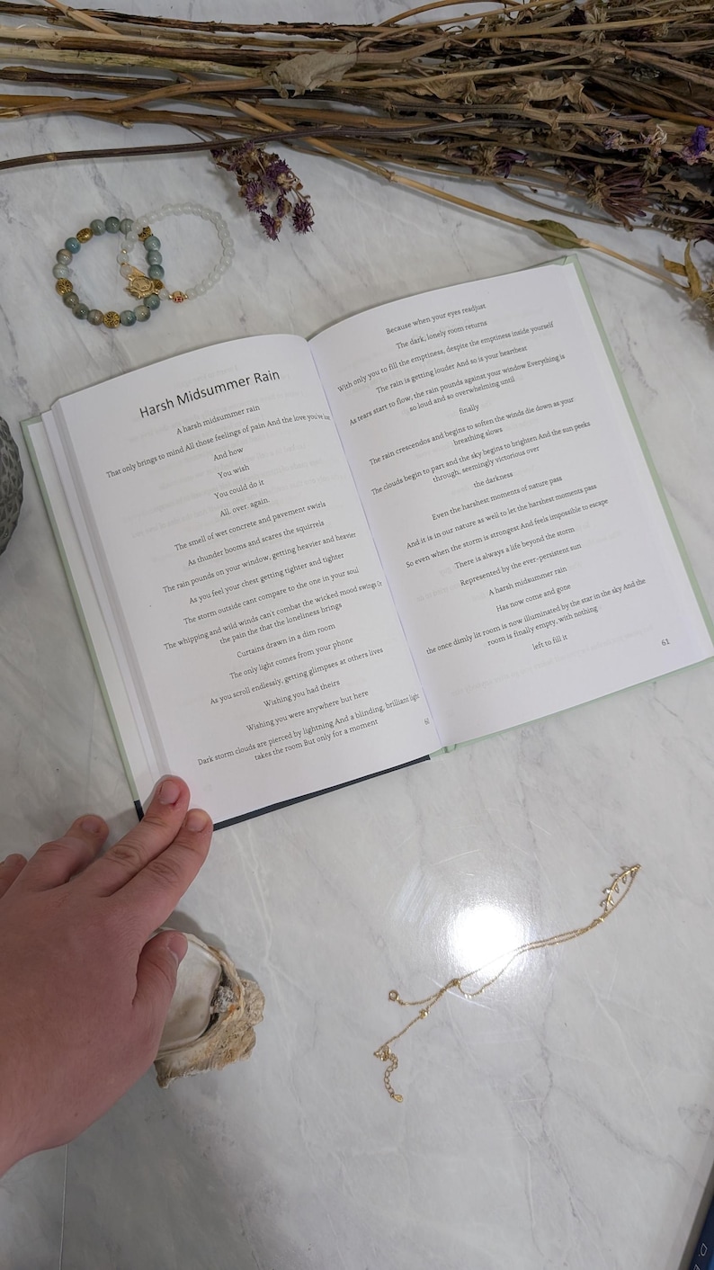 Pu&ograve; includere: Un libro aperto con il titolo "Harsh Midsummer Rain" e testo sulle pagine. Due braccialetti di perline e una collana d'oro sono accanto al libro. Fiori e steli secchi sono sparsi intorno al libro.