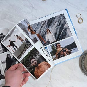 May include: An open photo album with multiple images, including black and white photos and color photos. The album is held open by a hand. The photos show people in various settings. Gold rings and a decorative plate are on the marble surface.