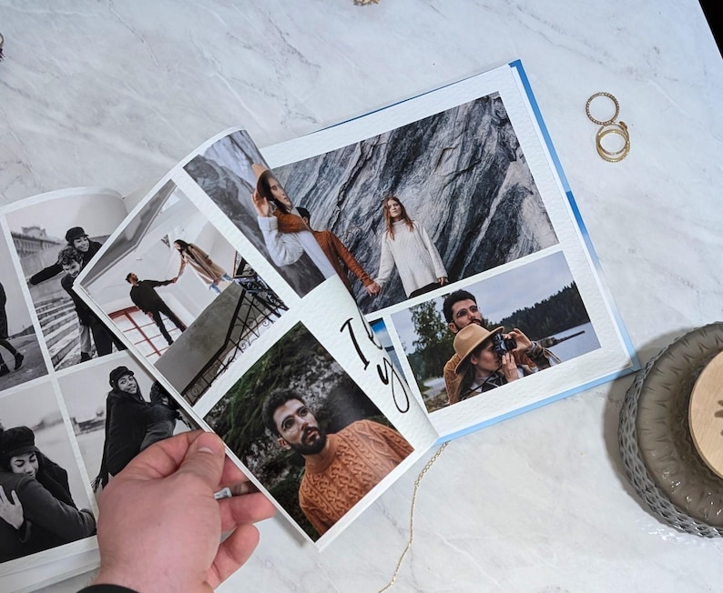 May include: An open photo album with multiple images, including black and white photos and color photos. The album is held open by a hand. The photos show people in various settings. Gold rings and a decorative plate are on the marble surface.