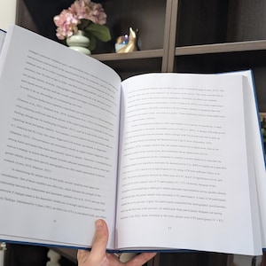 Puede incluir: Una persona sostiene un libro abierto con texto en las p&aacute;ginas. El libro tiene una cubierta azul. Un estante con objetos decorativos se encuentra en el fondo.