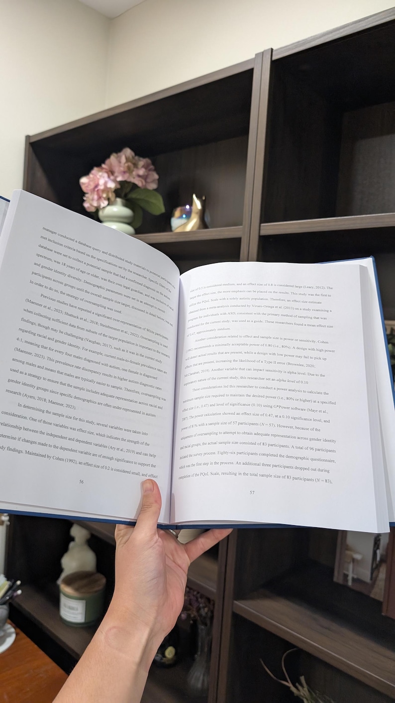 Puede incluir: Una persona sostiene un libro abierto con texto en las p&aacute;ginas. El libro tiene una cubierta azul. Un estante con objetos decorativos se encuentra en el fondo.