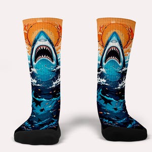 Puede incluir: Un par de calcetines coloridos con un gráfico de la cabeza de un tiburón rompiendo el agua. Los calcetines son azules, naranjas y blancos con un fondo de puesta de sol.