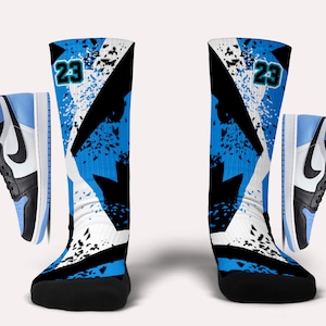 Puede incluir: Un par de calcetines deportivos azules, negros y blancos con un diseño de salpicaduras y el número "23" en cada calcetín. Los calcetines se combinan con una zapatilla de baloncesto azul claro y negra.