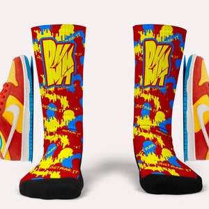 Puede incluir: Un par de calcetines rojos, amarillos y azules con el texto "Just Dunk It" y un logotipo de baloncesto. Los calcetines se combinan con un par de zapatillas de deporte naranja y rojas con un swoosh amarillo.