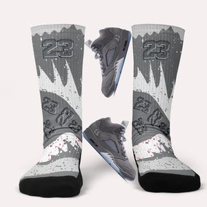 May include: Grey and white knee-high socks with a basketball theme, featuring the number 23 and a splatter design. Two grey trainers are also in the image, one above and one below the socks. The socks have black toes and heels.