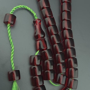 May include: A string of deep red, translucent, barrel-shaped beads threaded on a bright green cord. The prayer beads feature a larger, ornate central bead and a single bead at the end, finished with a small, matching green tassel.