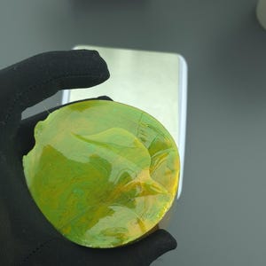 May include: A circular, abstract artwork in shades of green and yellow. The piece is held by a gloved hand, highlighting its texture and colour variations. It appears to be a unique decorative item.