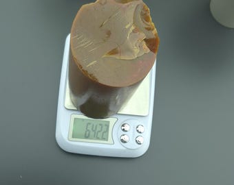 May include: A brown, irregularly shaped object is placed on a digital scale, displaying a weight of 64.22. The scale is white with silver buttons. Decorative plants are in the background.