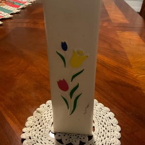 May include: A tall, off-white vase with a rectangular shape. The vase is decorated with three colorful tulip designs in blue, yellow, and red. The vase is sitting on a white, crocheted doily.