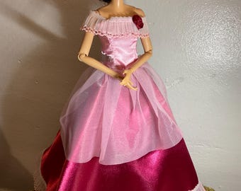 May include: A doll wearing a pink and red gown with a ruffled neckline and a red rose accent. The doll has dark hair and is posed with its arms at its sides. The dress has a layered skirt with a sheer overlay.