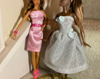 May include: Two fashion dolls wearing formal dresses. One doll is wearing a pink satin dress with a black belt and pink heels. The other doll is wearing a silver sequined dress with white flowers and blue heels.