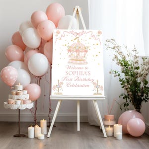 May include: A white easel holds a sign with a carousel illustration and the text "Welcome to Sophia's First Birthday Celebration." The scene is decorated with pink and white balloons, candles, and flowers. The sign is the centrepiece of a birthday party.