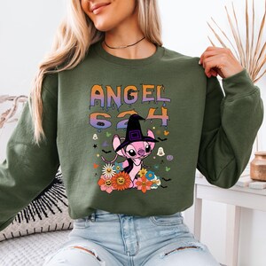 May include: Olive green sweatshirt featuring a graphic of the character Angel 624 wearing a witch hat, surrounded by flowers and Halloween-themed icons. The text "ANGEL 624" is printed above the character in a gradient color scheme.