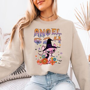 May include: Beige sweatshirt featuring a cartoon character wearing a witch hat, surrounded by flowers and Halloween-themed graphics. The text "ANGEL 624" is printed above the character in a colorful, dripping font. The sweatshirt is a neutral color.