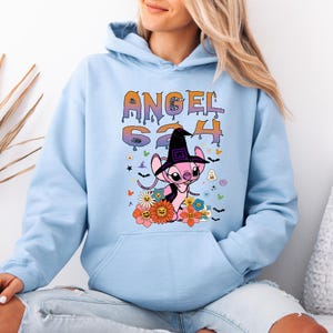 May include: Light blue hoodie featuring a graphic of a pink cartoon character wearing a witch hat, surrounded by flowers and Halloween-themed elements. The word "ANGEL" and the numbers "624" are displayed in a colorful, dripping font.