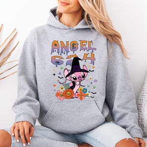 May include: A light gray hoodie featuring a graphic of a pink alien character wearing a witch hat, surrounded by flowers and bats. The text "ANGEL 624" is printed above the character in orange and purple. The hoodie has a drawstring hood.