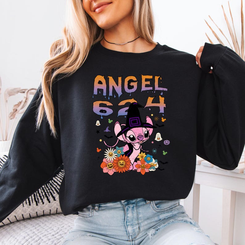 May include: Black sweatshirt featuring a pink cartoon character wearing a witch hat, surrounded by flowers and Halloween-themed graphics. The text "ANGEL 624" is displayed in colorful lettering above the character. A fun, festive design.