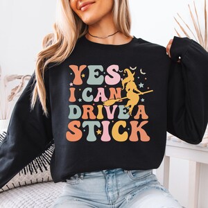 May include: Black sweatshirt with the text "YES I CAN DRIVE A STICK" in retro colors. A witch on a broom is also featured. The design includes stars and bats, perfect for Halloween.