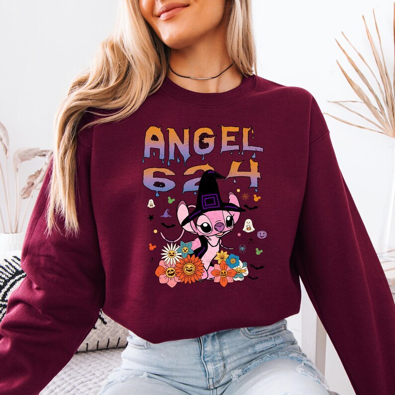 May include: Burgundy sweatshirt with the text "ANGEL 624" in orange and purple dripping letters. A pink cartoon character wearing a witch hat is surrounded by flowers, bats, and ghosts.