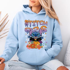 May include: Light blue hoodie featuring a graphic of Stitch from Lilo & Stitch dressed as a vampire, with the text "Stitch 626". The design includes a pumpkin, flowers, and Halloween-themed elements.