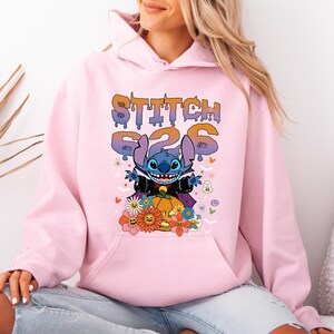 May include: A light pink hoodie featuring a graphic of Stitch from Lilo & Stitch. The graphic includes the text "STITCH 626" in a dripping font, Stitch in a Halloween costume, a pumpkin, and flowers.