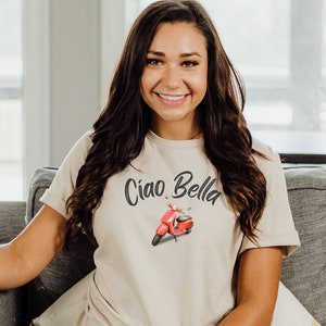 May include: A woman wearing a beige t-shirt with the text "Ciao Bella" and a red scooter graphic. She is sitting on a gray couch with a white pillow and a yellow striped pillow.