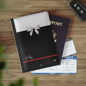 May include: Black passport holder with a white bow and the name "Kathy" printed in red. The holder has a red heart with a plane and a dotted line. The holder is next to a blue passport and two airline boarding passes.
